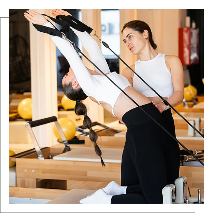 A woman assists another woman performing a backbend exercise on a Pilates reformer in a brightly lit studio.