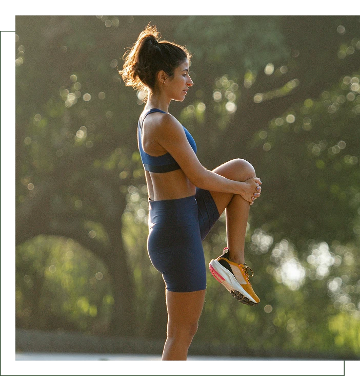 A woman in athletic wear stands outdoors, holding one knee up to her chest in a stretching pose, with sunlight and trees in the background.