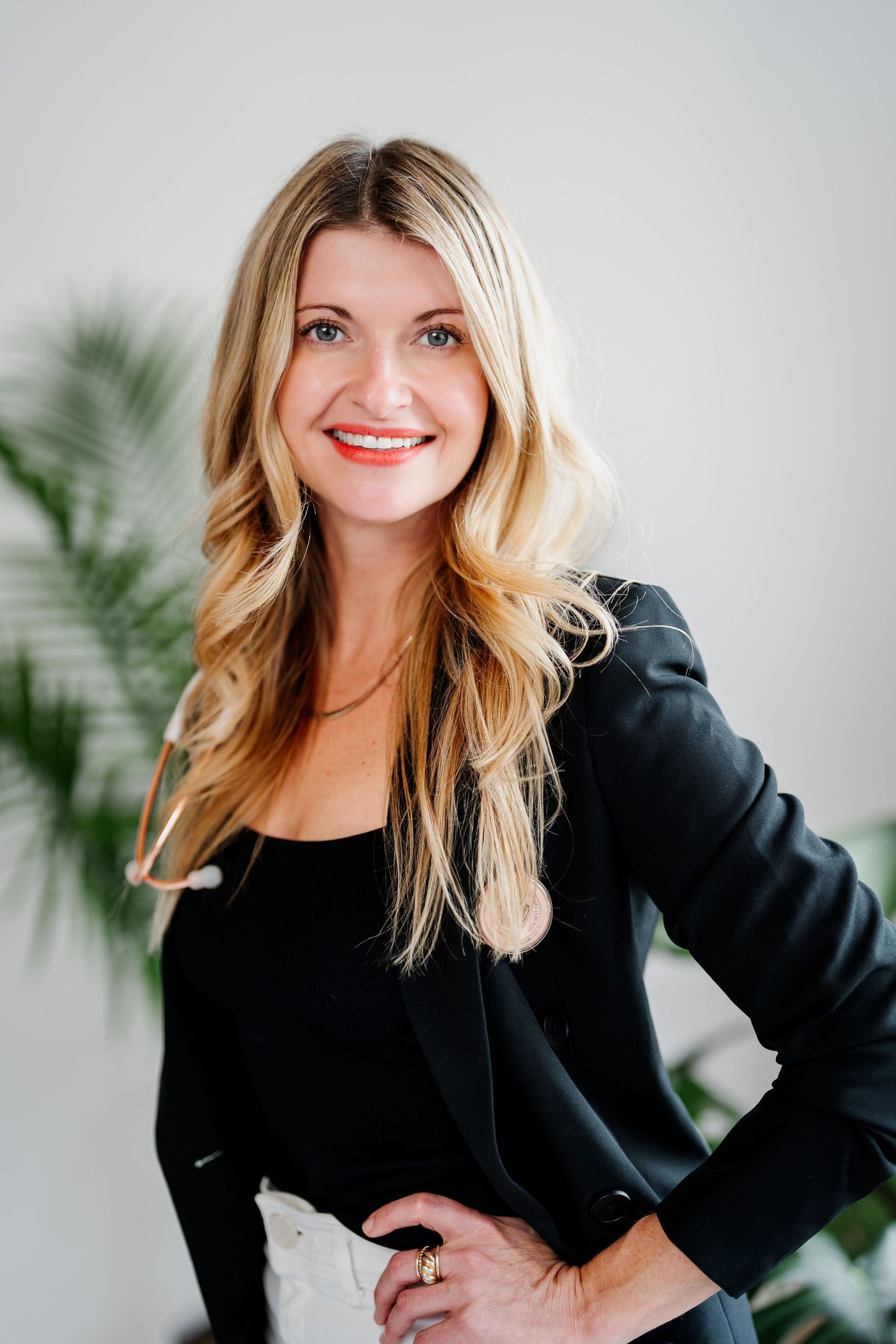 Woman with long blonde hair wearing a black blazer, black top, and white pants, standing indoors with a hand on her hip and smiling at the camera. A leafy plant is in the background.